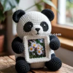 A crocheted panda sitting on a wooden stool, holding a small photo frame with a picture of white daisies.