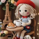 Crochet doll wearing a red beret with a striped shirt, sitting at a small table with a coffee and a croissant. A small crochet dog is beside the table, and an Eiffel Tower model and flowers are in the background. A "Bonjour" sign is displayed above.