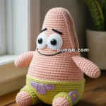 A knitted plush toy resembling a pink starfish character with large eyes and a smiling face, dressed in green shorts with a purple flower, sitting on a wooden table next to a plant.