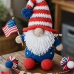 A crocheted gnome wearing a red, white, and blue striped hat holds a small American flag. It has a white beard, blue body with star patterns, and red feet, sitting on a wooden table adorned with decorative stars and the letters 'USA' in crochet. Twinkling lights are visible in the blurred background.