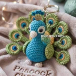 A crocheted peacock keychain with vibrant blue and green colors sits on a soft pink fabric embroidered with the words 'Peacock Charm.' The keychain features intricate detailing with small round eyes and a stylized tail.