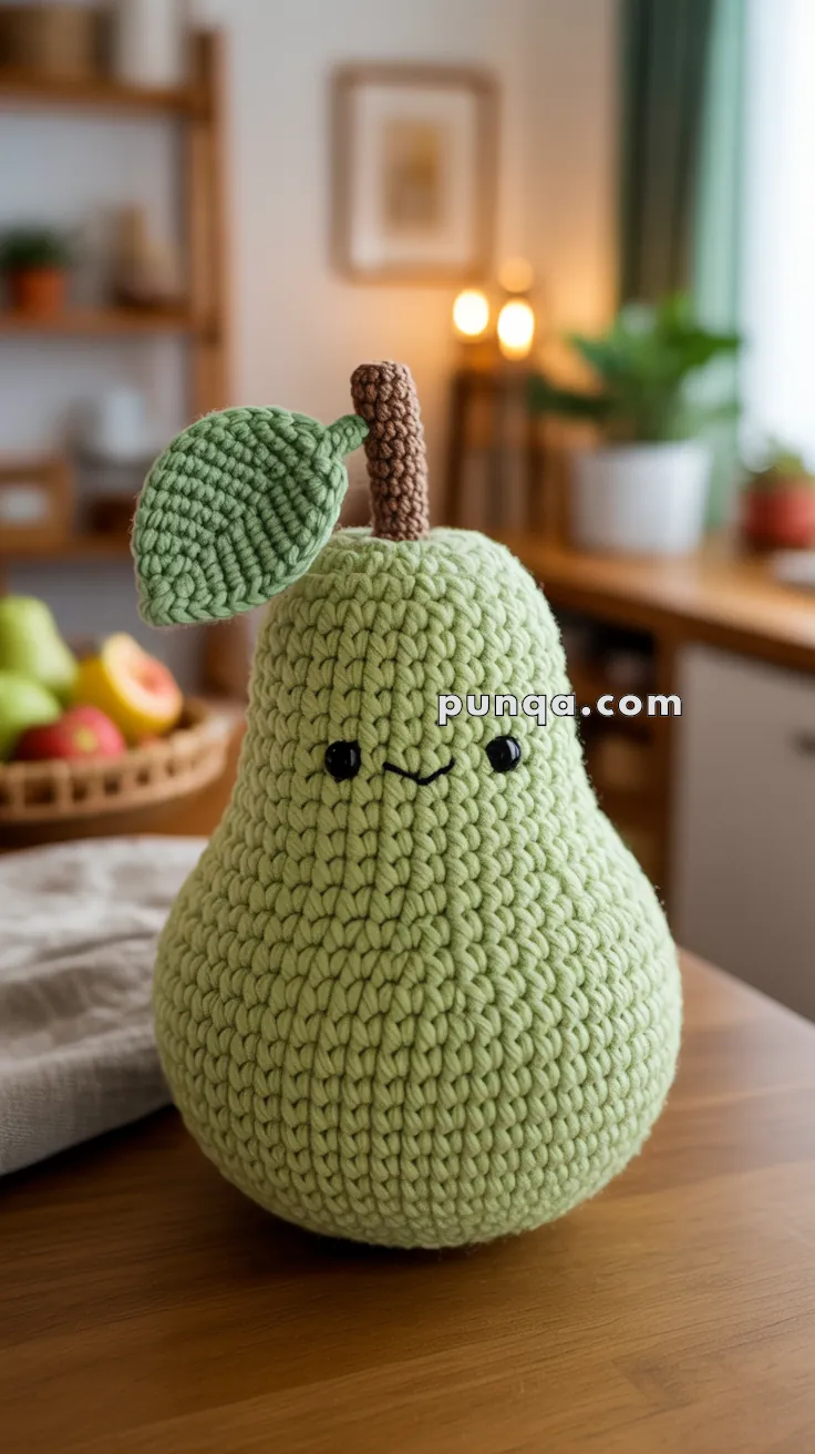 A crocheted pear-shaped plush toy with a smiling face, featuring green yarn for the body and leaf, and brown yarn for the stem, sitting on a wooden table in a cozy kitchen setting.