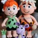 Crocheted dolls depicting two characters with prehistoric attire and a purple dinosaur, crafted with detailed stitching and placed on a knitted mat. Small crochet rocks and a plant on a stool complete the scene.
