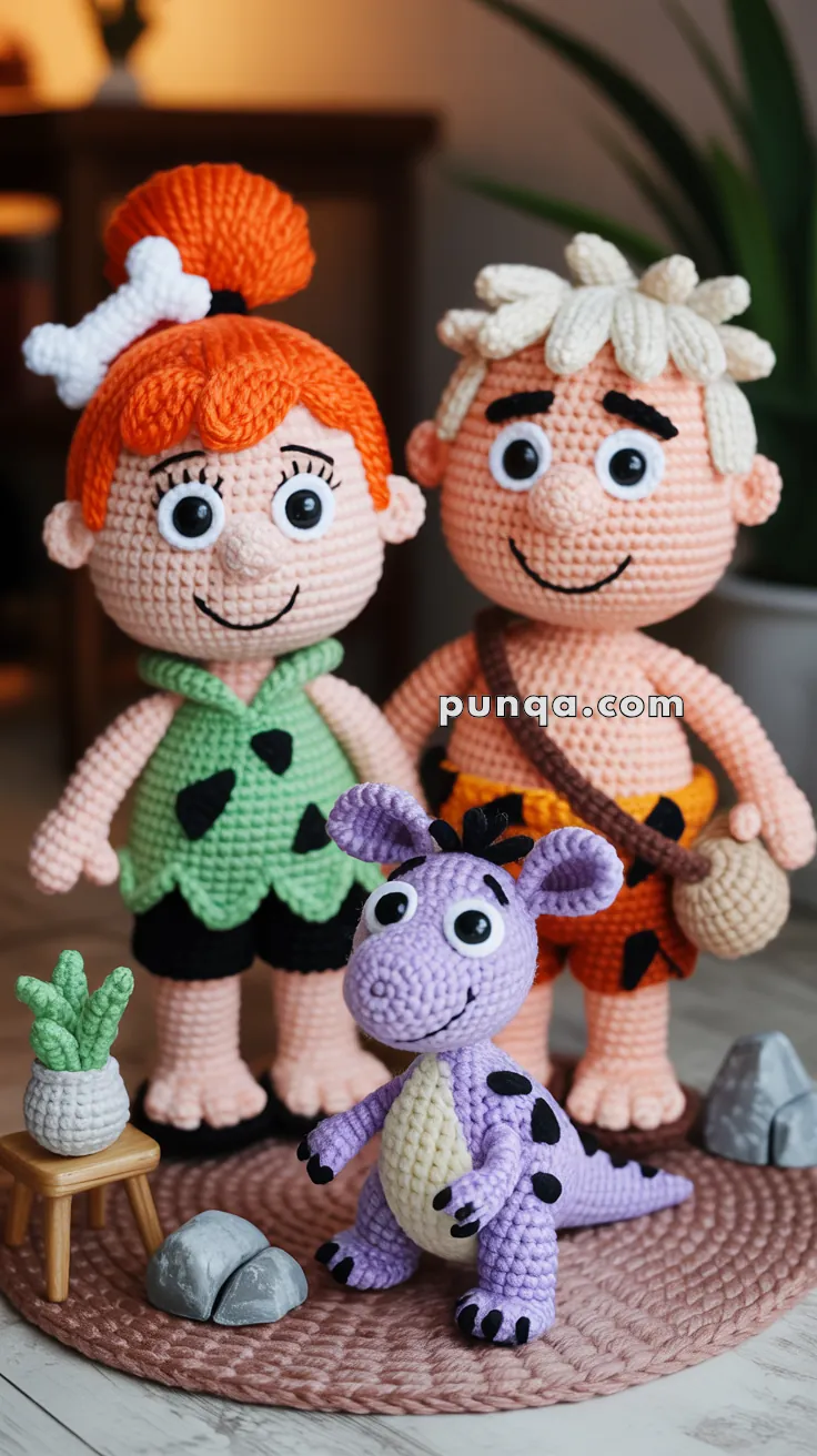 Crocheted dolls depicting two characters with prehistoric attire and a purple dinosaur, crafted with detailed stitching and placed on a knitted mat. Small crochet rocks and a plant on a stool complete the scene.