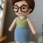A crocheted doll with brown hair, large round glasses, a green top, and a blue skirt, standing on a wooden surface.