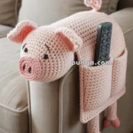 A crocheted pig-shaped armrest cover with pocket sits on the arm of a beige couch, holding a remote control.