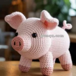 A handmade crochet pig toy with pink yarn and black button eyes, placed on a wooden table in a cozy room setting with blurred lights and plants in the background.