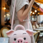 A woman holding a cute crochet handbag designed to look like a pig's face, featuring a round shape, prominent snout, and smiling expression, with a background of an outdoor cafe setting.