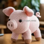 A crocheted pink pig with black eyes and a curly tail sits on a wooden table, with a soft-focus room interior in the background.