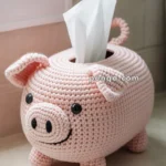 A crocheted pink pig-shaped tissue box cover with tissues protruding from the top, placed on a bathroom countertop near a window.