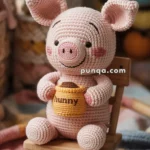 A crocheted piglet toy with a smiling face and pink cheeks, holding a small yellow crocheted pot labeled "hunny," seated on a wooden chair.