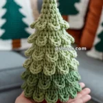 A hand holding a crocheted green Christmas tree with layered, scalloped edges, in front of a sofa with festive cushions depicting trees.