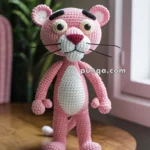 A crocheted pink panther toy with large eyes and a white belly, standing on a wooden table near a window with sunlight streaming in.