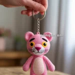A hand holding a crocheted pink panther keychain, featuring a cartoonish face with yellow eyes and a slight smile, set against an indoor background.