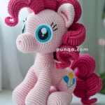 Crocheted toy pony with pink yarn and curly mane, blue eyes, and colorful balloon designs on its flank, placed on a white table.