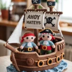 A crocheted pirate ship with two pirate dolls aboard, featuring a sail that reads 'AHOY MATEYS' and a skull design, placed on a wooden table.