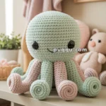 A cute crocheted octopus plush in shades of mint green and pink sits on a shelf, surrounded by other soft toys and baby blankets in a nursery setting.