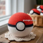 A knitted Poké Ball placed on a wooden table with crochet doilies nearby, and more knitted balls in a basket in the background.