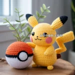 A crocheted Pikachu doll standing next to a crocheted Poké Ball on a wooden table, with a green plant in the background.