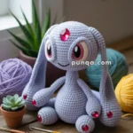 A handmade crocheted plush toy with large eyes and long ears, embellished with red gems, is sitting next to balls of yarn and crochet hooks on a wooden surface. A small potted succulent and other yarn supplies are also visible in the background.