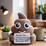 A crocheted toy shaped like a smiling poop emoji holds a sign with an encouraging message, sitting on a wooden table with a plant and a steaming mug nearby.
