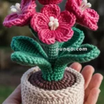 A hand holding a crocheted plant with pink flowers and green leaves in a beige crocheted pot.