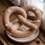A knitted pretzel with white yarn dots sits on a decorative plate that reads "BAKED WITH LOVE" on a wooden surface, next to a window with sheer curtains.