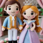 Crocheted dolls of a prince and princess, with detailed attire and crowns, standing on a decorative pink surface with crocheted roses.