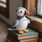 A knitted puffin toy with orange beak and feet stands on a stack of vintage books, placed on a wooden windowsill.