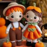 Crocheted dolls of a boy and a girl standing together on a round orange mat, surrounded by small crocheted pumpkins and leaves. The boy is wearing a straw hat and brown overalls, and the girl is in an orange dress with a pumpkin-themed hat. The background includes Halloween decorations like a jack-o'-lantern and soft glowing lights.