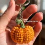 A hand holding a keychain with a crocheted orange pumpkin attached to it, featuring a green stem, against a softly blurred indoor background.