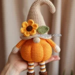 A handmade crocheted gnome with a beige curly hat, pumpkin orange body, and striped brown and white legs, held in a person's hand.