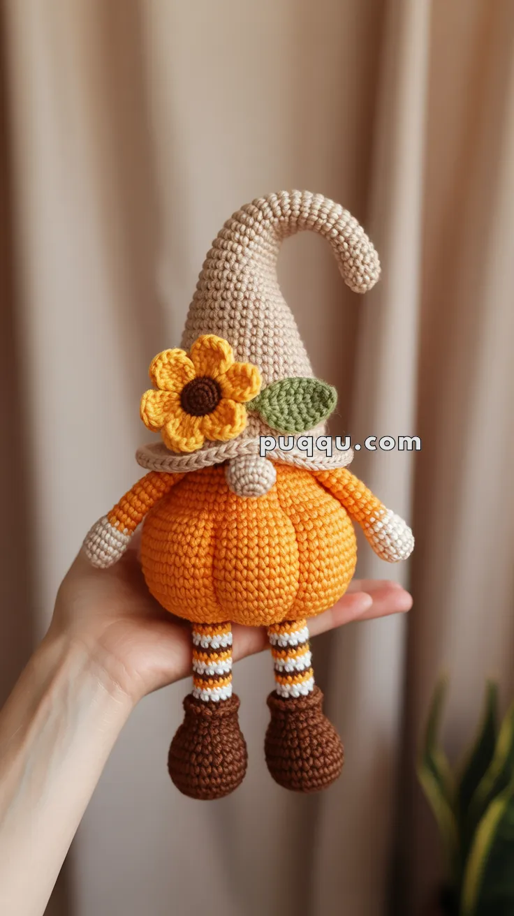 A handmade crocheted gnome with a beige curly hat, pumpkin orange body, and striped brown and white legs, held in a person's hand.