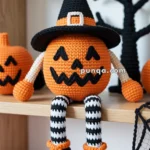 A handcrafted crochet pumpkin doll wearing a black witch hat and sitting on a shelf. It has striped black-and-white legs and black shoes with orange laces. Decor elements include another pumpkin, a spider web, and a tree silhouette in the background.