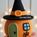 A crocheted decoration shaped like a pumpkin, featuring a green door, a blue and black window, and a black witch hat with an orange band, held in a person's hand against a blurred background with lights.