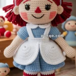 A crocheted doll with red yarn hair, a blue dress, and a white apron, featuring a smiling face with large eyes and a red nose, standing in a room with other colorful crocheted figures in the background.