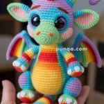 Colorful crocheted dragon plush toy with rainbow stripes and large black eyes, held in a person's hand.