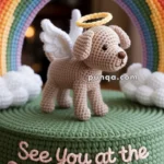 Crocheted sculpture of a dog with angel wings and a halo, standing on a green base with the text 'See You at the Rainbow Bridge', set against a rainbow backdrop.