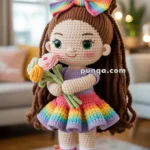 Crocheted doll with a large rainbow bow, wearing a rainbow dress and holding a bouquet of crocheted flowers, standing on a wooden surface in a cozy indoor setting.