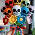A colorful crochet bouquet featuring skull heads in various colors, each with black eye sockets and nose details. The stems and leaves are also crocheted, tied with a multicolored bow, and surrounded by multicolored crochet flowers. The background shows a blurred room with rainbow decorations.