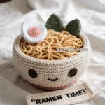A crochet bowl of ramen with a cute face, featuring noodles, a crocheted egg, and green leaves, placed on a textured fabric background. A crocheted label with the text 'RAMEN TIME!' is in front of the bowl.