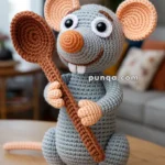 A crocheted gray and peach-colored mouse plush toy sitting on a wooden table, holding a brown crochet spoon, with a blurred living room in the background.