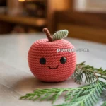 A crocheted red apple with black button eyes, a smile, and a green leaf on a brown stem, set on a wooden table with a sprig of greenery beside it.