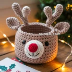 A crocheted deer basket with antlers and a red nose, accompanied by a "Merry Christmas" card, is placed on a wooden table adorned with Christmas lights, with a blurry Christmas tree in the background.