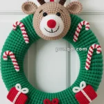 A crocheted Christmas wreath featuring a reindeer head with a red nose, candy canes, and small gift boxes, hanging on a door with a red ribbon.