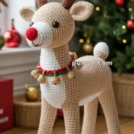 A handmade crocheted reindeer toy with beige and white yarn, featuring a red nose and antlers. It is adorned with a festive bell collar and is placed in front of a Christmas tree with ornaments.