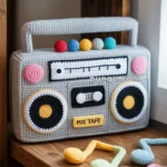 Crocheted boombox with colorful knobs and cassette tape design, labeled 'MIX TAPE,' displayed on a wooden shelf with crocheted musical notes nearby.