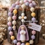A handmade crochet rosary with pastel-colored beads and a small crocheted figure of a praying person in a purple cloak, displayed on a wooden log slice with a tag reading 'Handmade with love.'