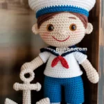 Crocheted sailor doll with a white sailor hat, blue shorts, white shirt, and red necktie, holding a crocheted anchor.
