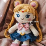 A crocheted Sailor Moon doll resembling a cartoon character with long blonde braids, wearing a sailor outfit in blue and white with a pink bow, and pink shoes, sitting on a soft pink fabric.